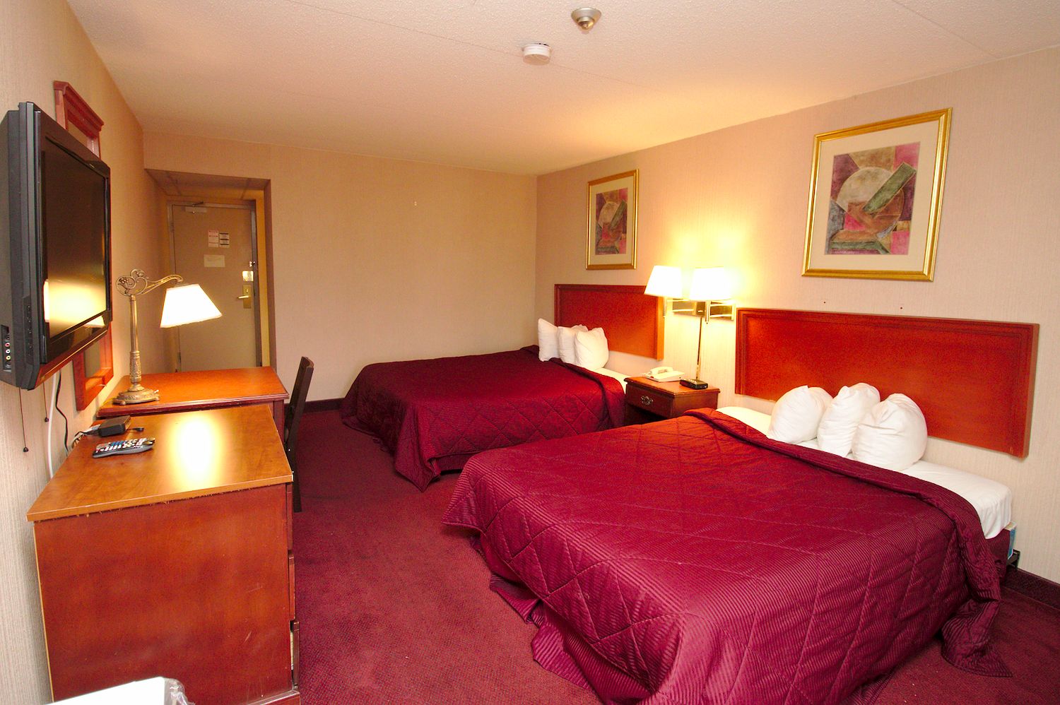 A hotel room with two beds, a desk with a lamp, a television, and artwork on the walls.