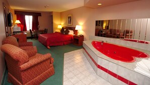 A hotel room with a large bed, a sitting area with two armchairs, a desk, a red bath tub, and a large mirror.