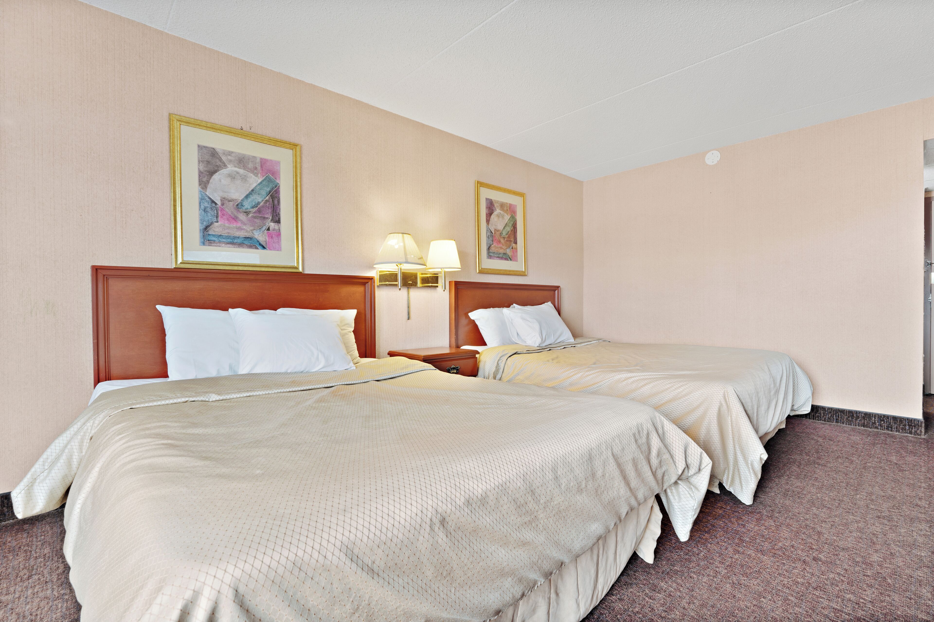 A hotel room with two beds, beige bedding, wooden headboards, and two wall-mounted lamps.