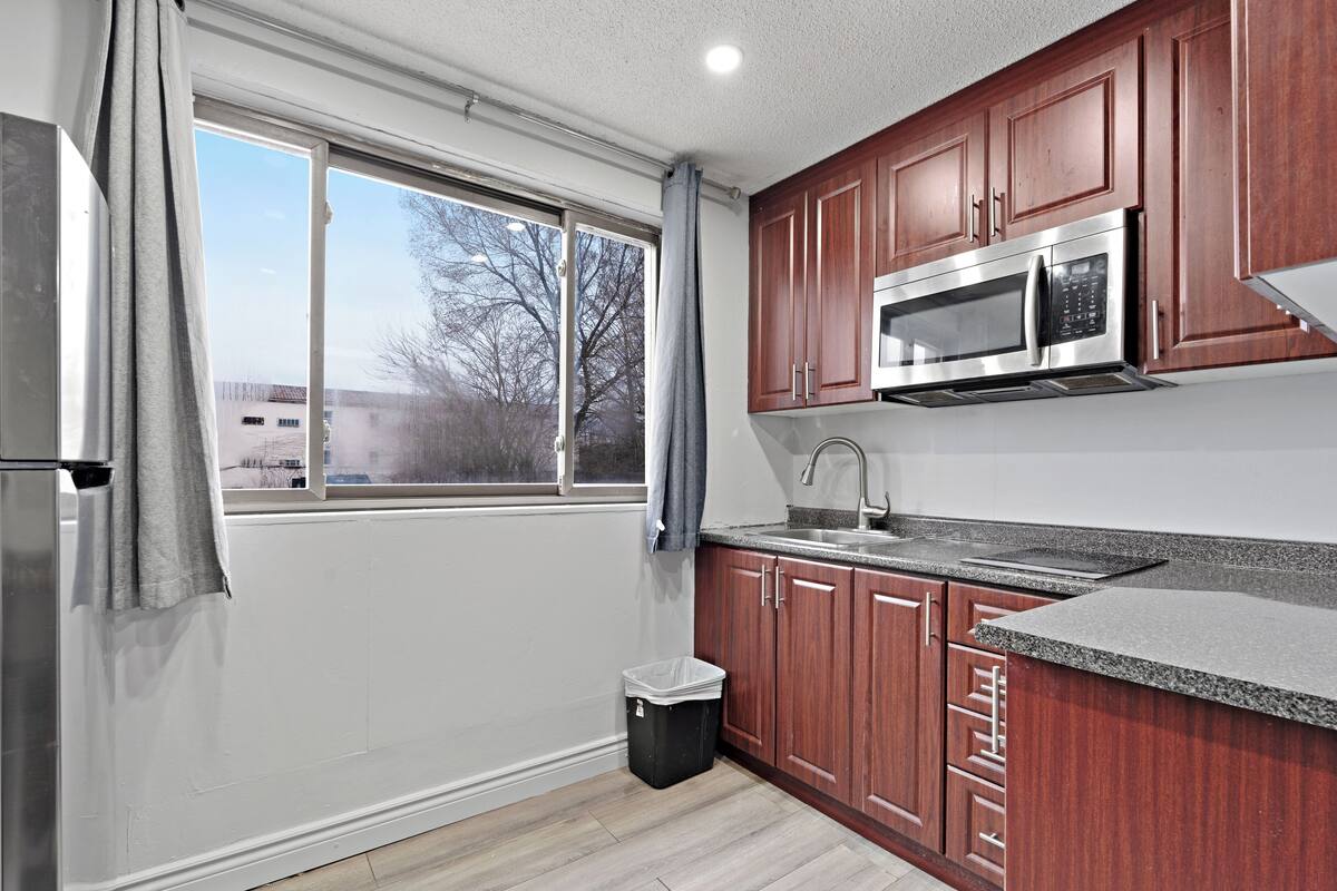 A kitchen with wooden cabinets, a stainless steel microwave, and a window with a view of trees and buildings.