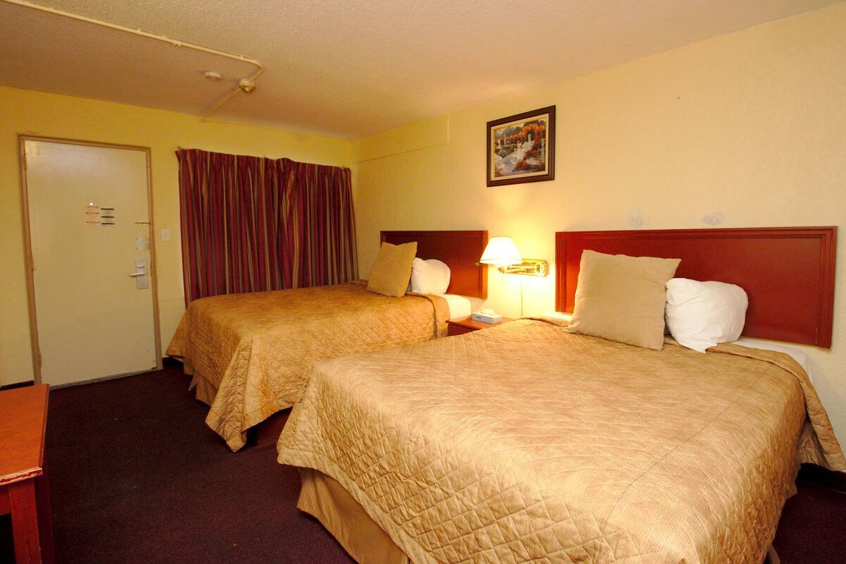 A hotel room with two beds, a door, a curtain, a nightstand, a lamp, and a painting on the wall.