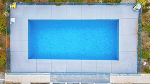 An aerial view of a rectangular swimming pool with a blue border, surrounded by a fence and some greenery.