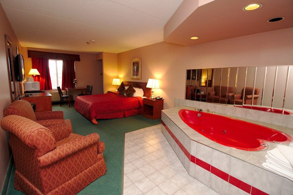 A hotel room with a large bed, a sitting area with two armchairs, a desk, a red-tiled bathtub, and a large mirror.