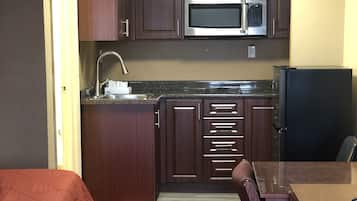 Une cuisine compacte avec des meubles de couleur bois foncé, un four à micro-ondes en acier inoxydable et un réfrigérateur. On y trouve également un petit coin repas avec une table et des chaises.