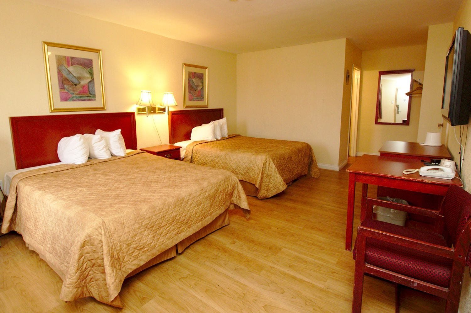 A hotel room with two beds, a desk, and a chair. There are two wall-mounted lamps and two framed pictures on the wall.