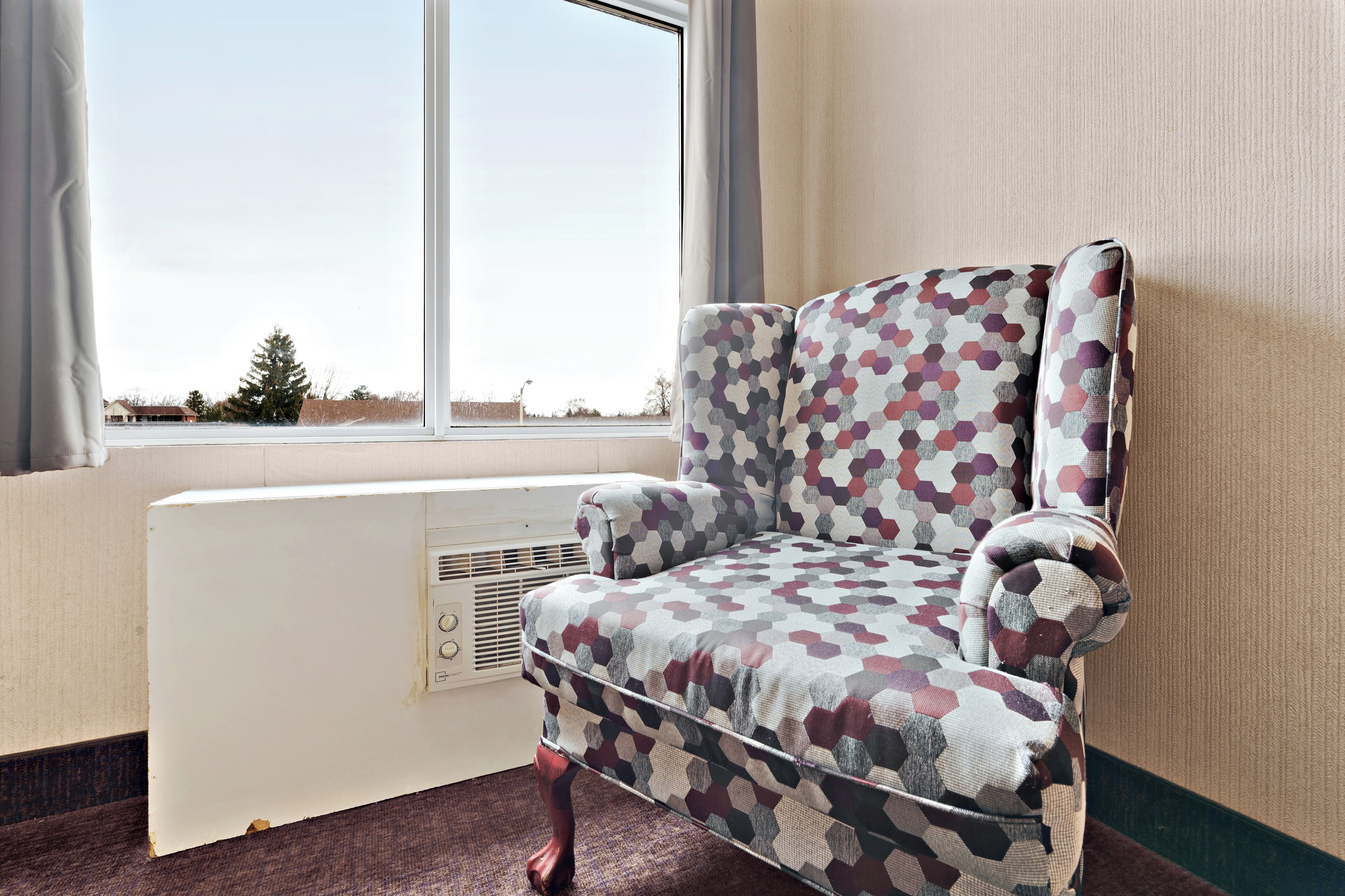 A room with a patterned armchair near a window, a radiator, and a view of a tree and buildings.