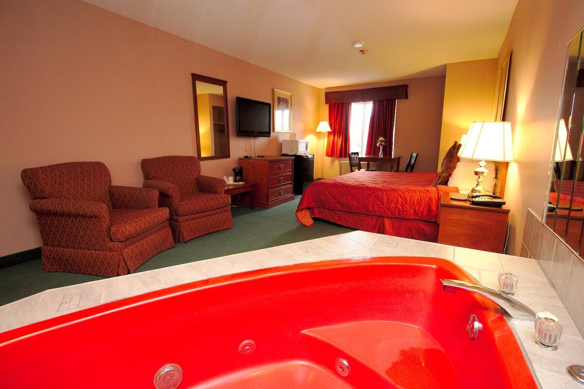 A hotel room with a red bathtub, two armchairs, a bed with a red bedspread, a television, a dresser, and a desk with a lamp.