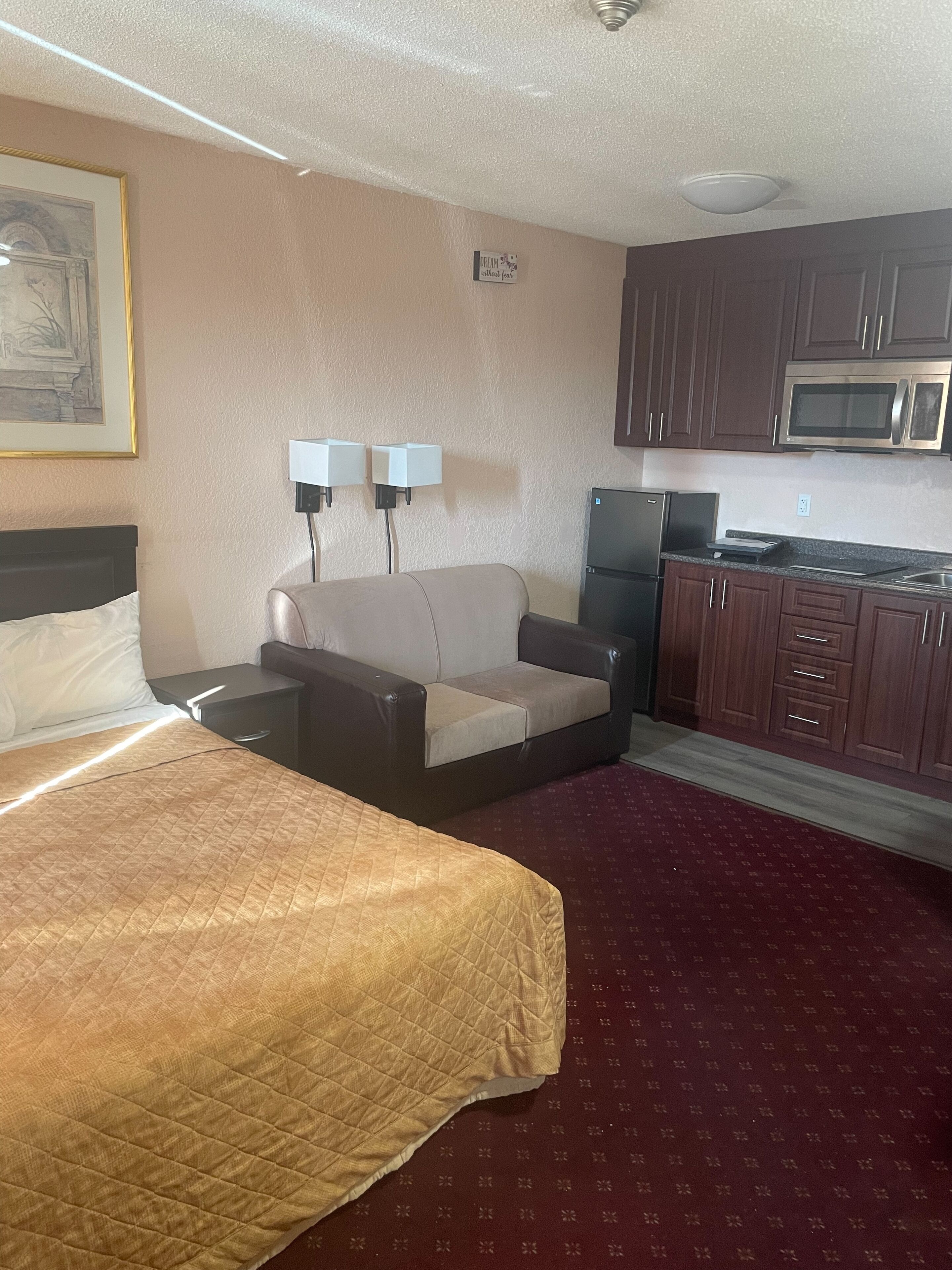 A hotel room with a bed, a sofa, a kitchenette with a microwave, and a painting on the wall.