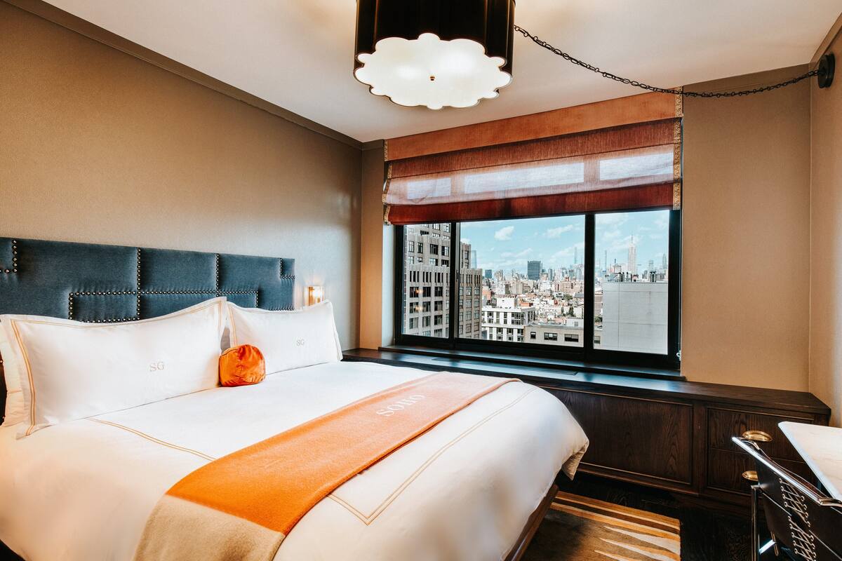 A hotel room with a large bed, a city view through the window, a headboard with a decorative panel, and a bedside table with a lamp.