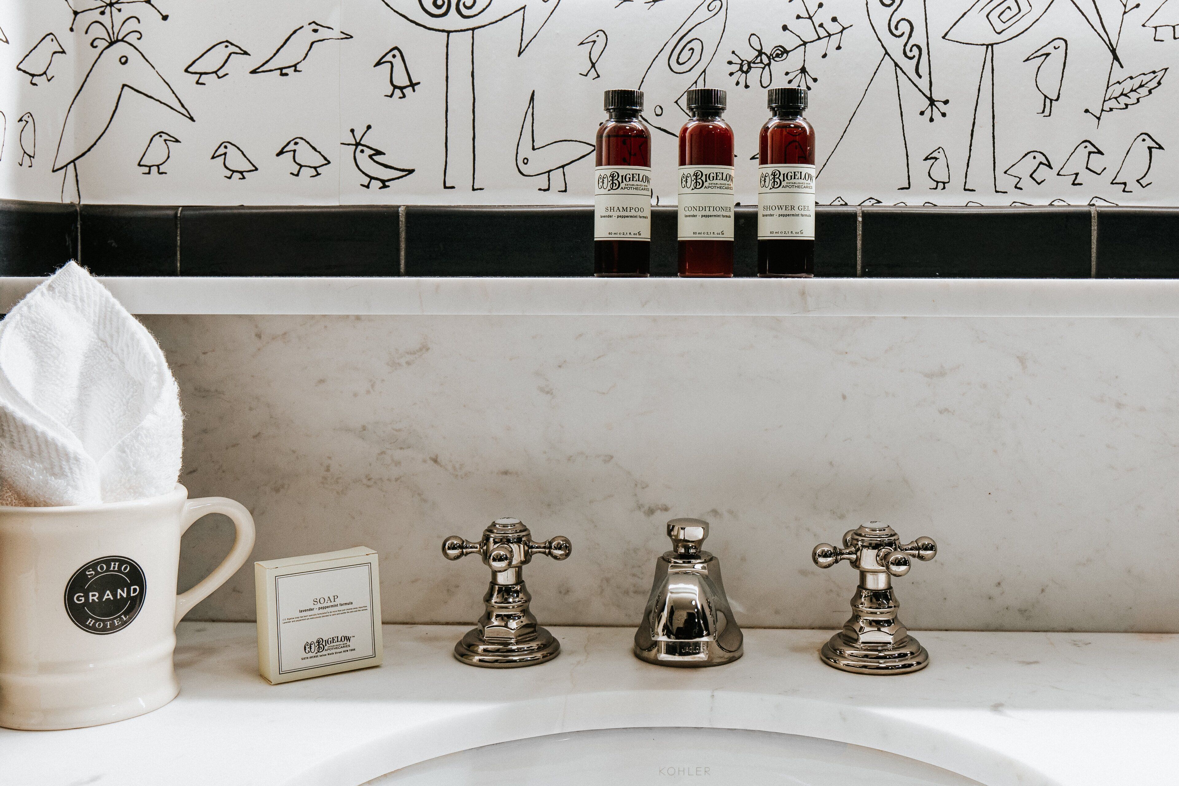 A bathroom with a white marble sink, a white mug labeled "SOHO GRAND HOTEL," and three bottles of "BIGELON" products.