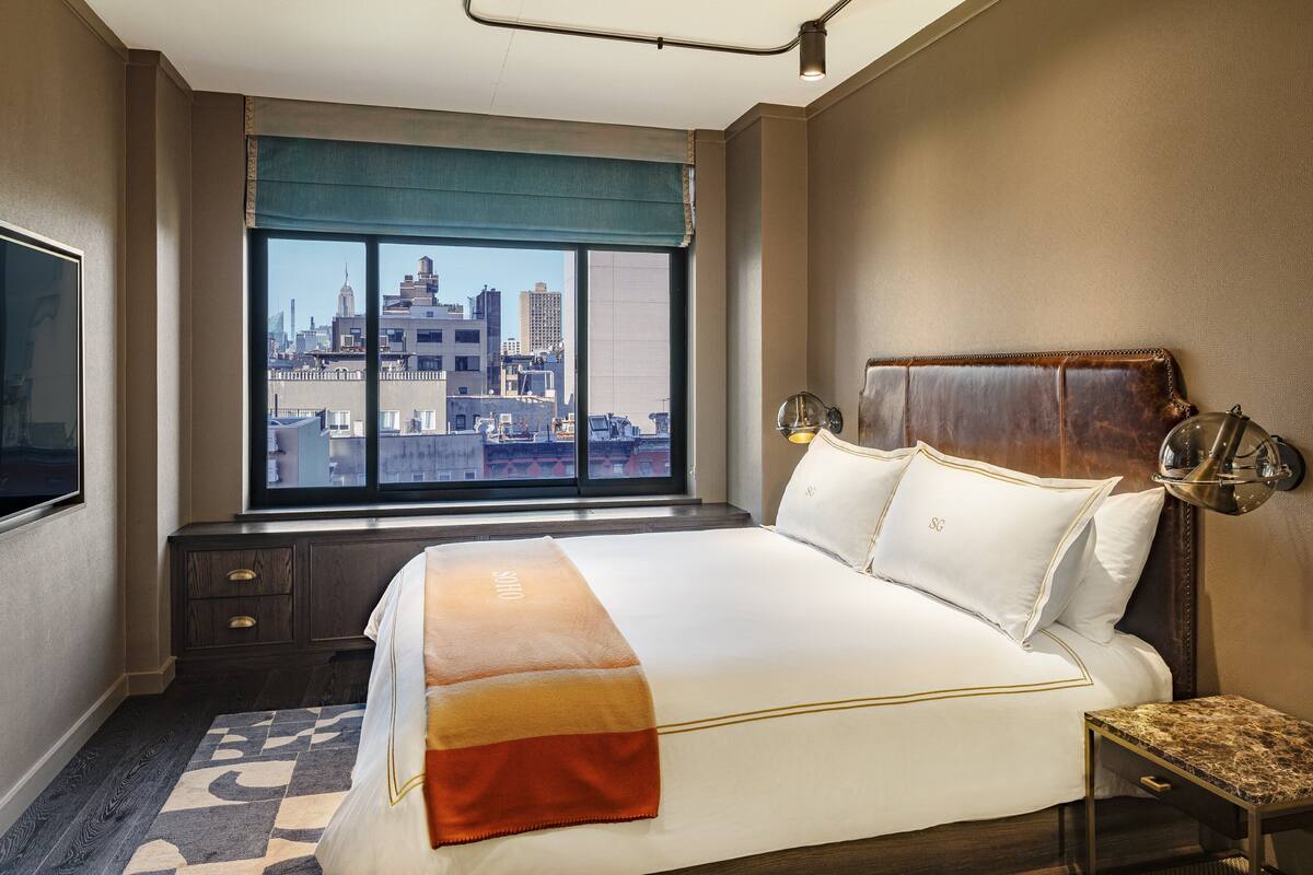 A hotel room with a large bed, a view of the cityscape, a wooden headboard, and a small table with a lamp.