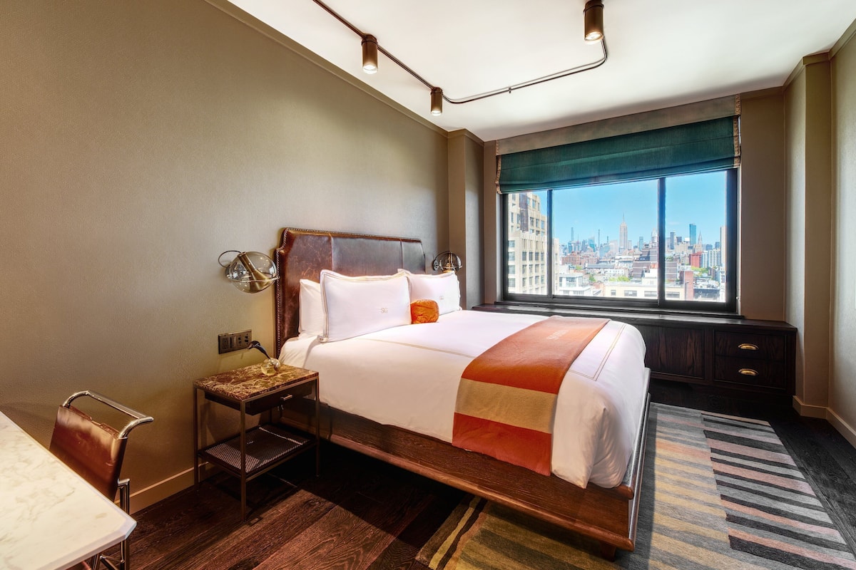 A hotel room with a large bed, a desk, a chair, a window with a city view, and a nightstand with a lamp.