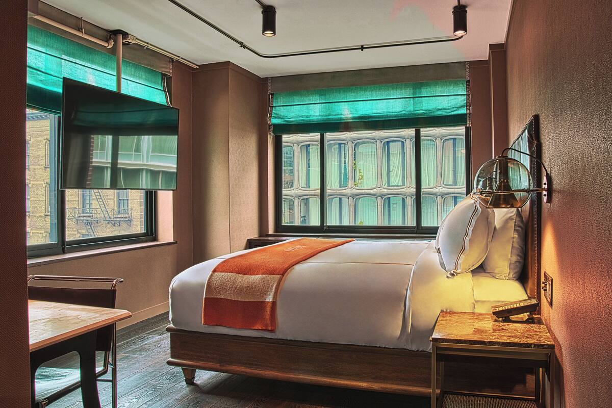 A hotel room with a large bed, a bedside table, a chair, a window with curtains, and a view of buildings outside.