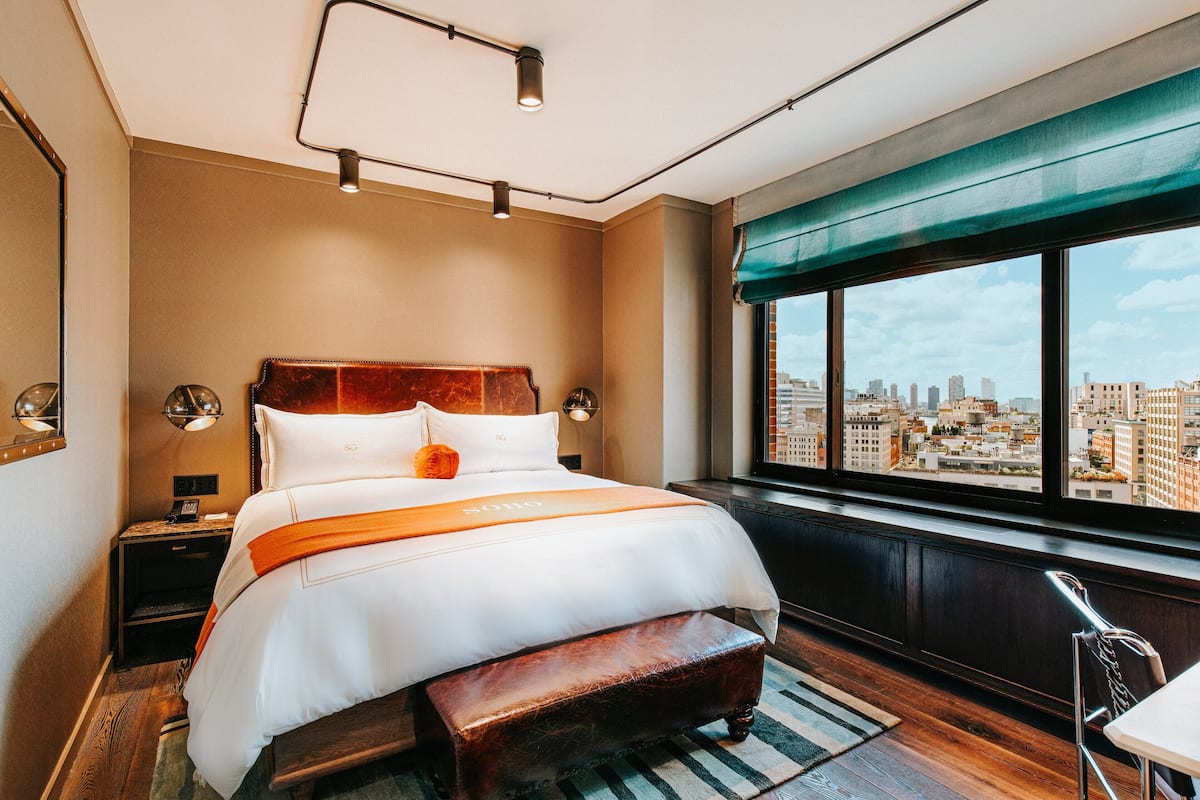 A modern hotel room with a large bed, a bench, a city view, and a cityscape visible through the window.