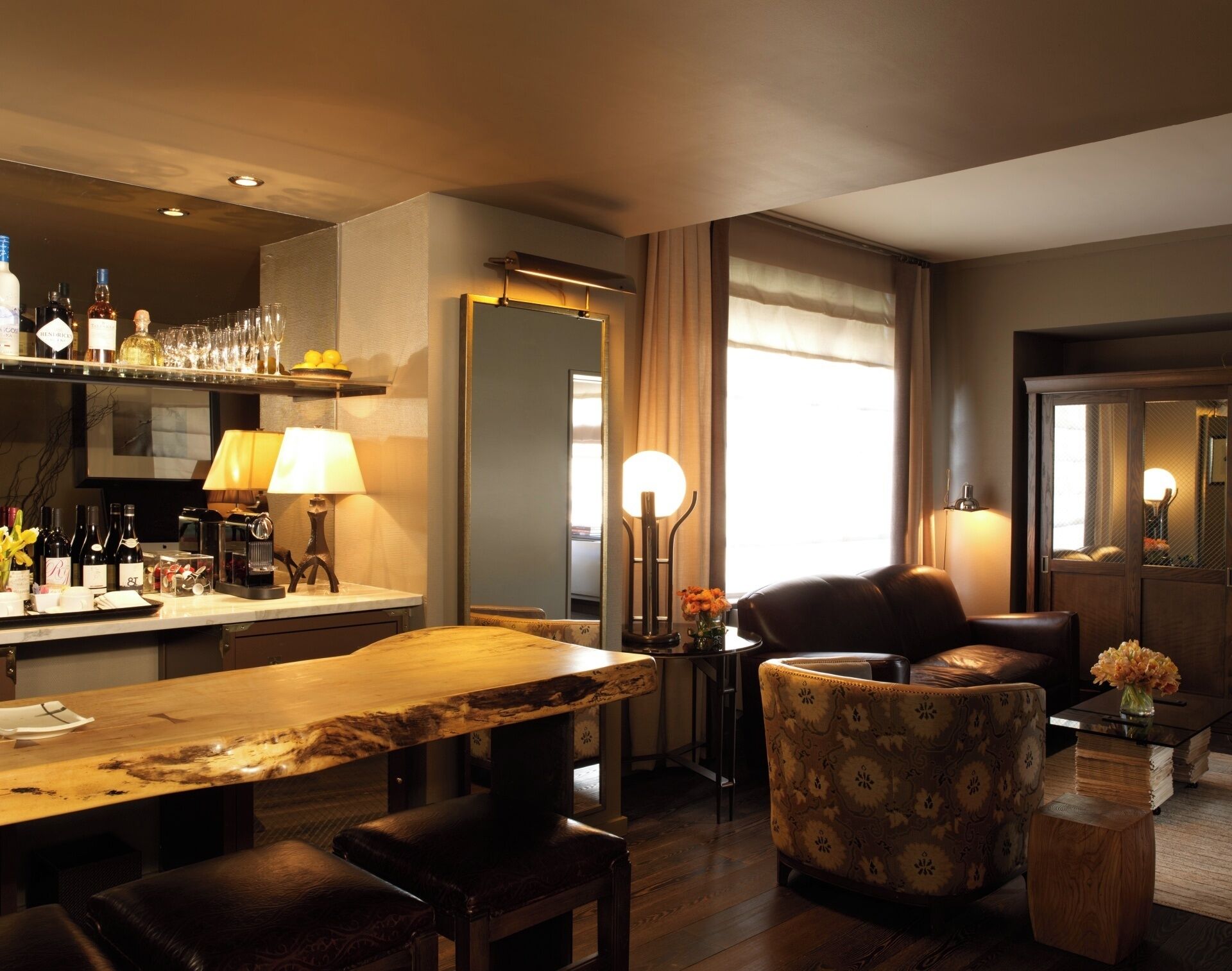 Habitación de hotel con una mesa de comedor de madera, sillas de cuero, una zona de bar con botellas y vasos, y un ventanal con cortinas.