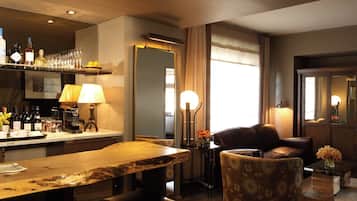 Habitación de hotel con una mesa de comedor de madera, sillas de cuero, una zona de bar con botellas y vasos, y un ventanal con cortinas.