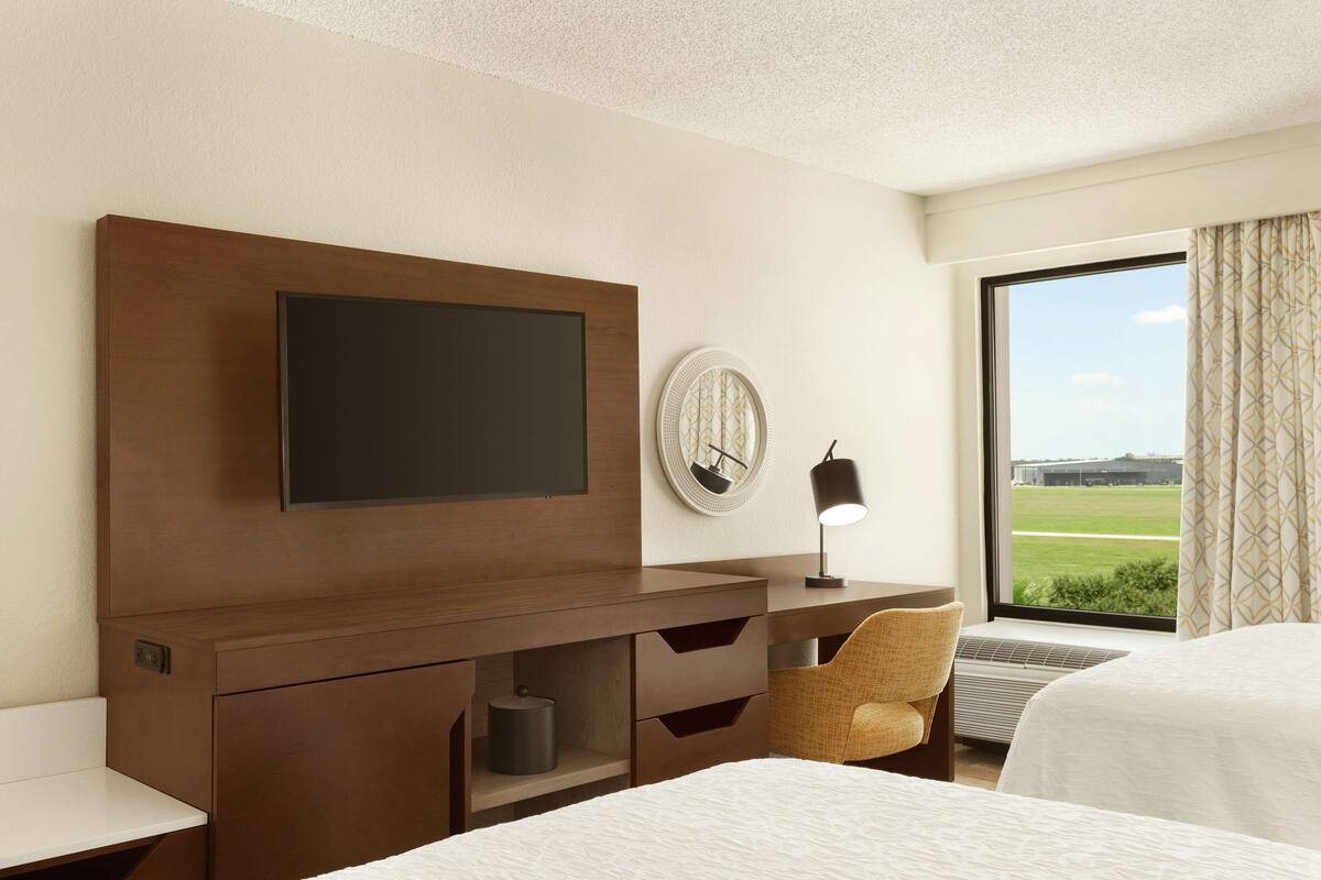Ein Hotelzimmer mit einem großen Flachbildfernseher an einem Holzkabinett, einem runden Spiegel, einem Schreibtisch mit Lampe, einem Sessel und einem Bett.