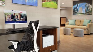 Un moderno vestíbulo de hotel con una mesa de recepción, un monitor de computadora mostrando una imagen promocional, una silla de malla negra, un sofá color beige con cojines y un gran cuadro de un paisaje urbano en la pared.