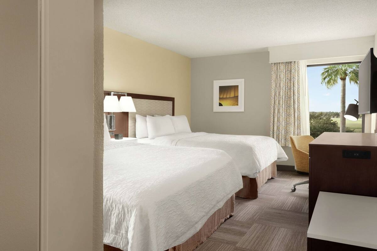 Ein Hotelzimmer mit zwei Betten, einem Schreibtisch, einem Sessel, einem Fernseher und einem Fenster mit Blick auf Palmen.