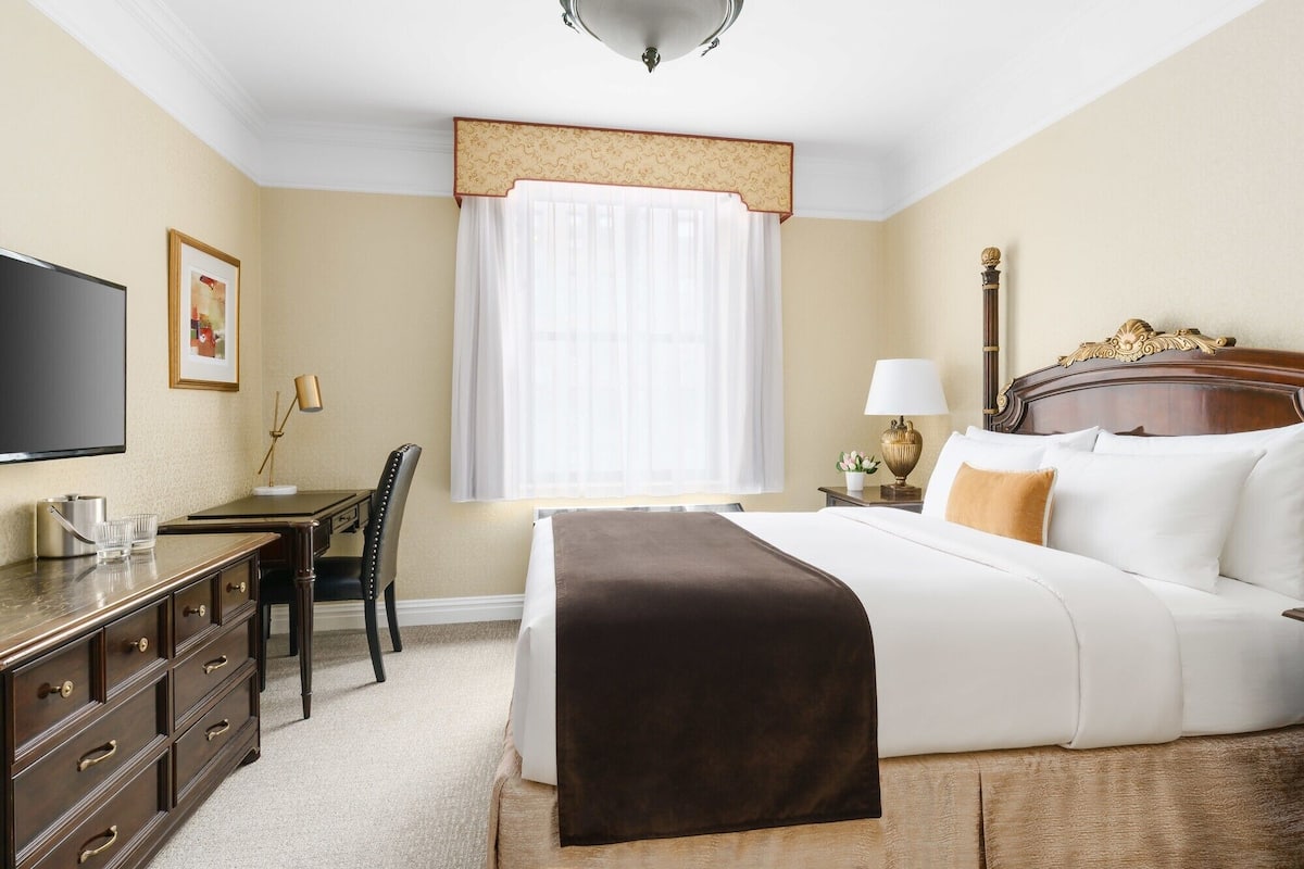A hotel room with a bed, a desk with a chair, a television, a lamp, and a window with curtains.