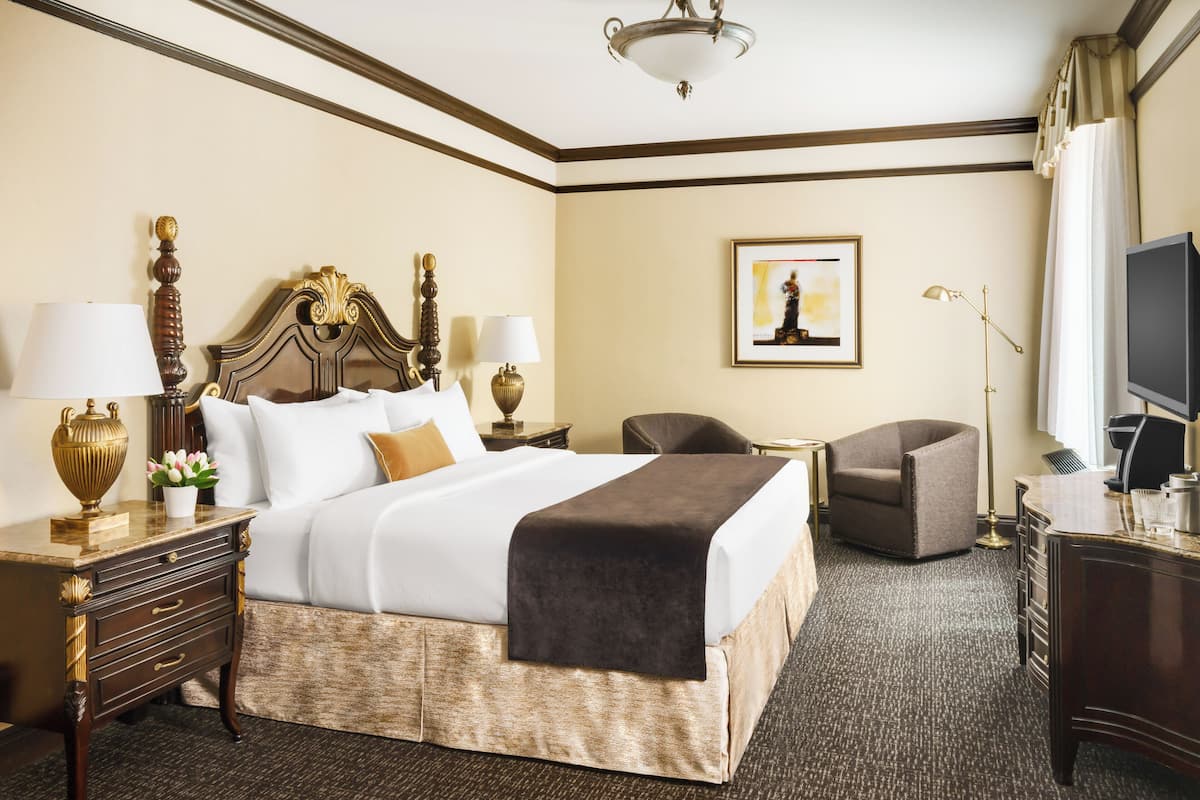A hotel room with a large bed, two bedside tables with lamps, a chair, a desk with a TV, and a framed picture on the wall.