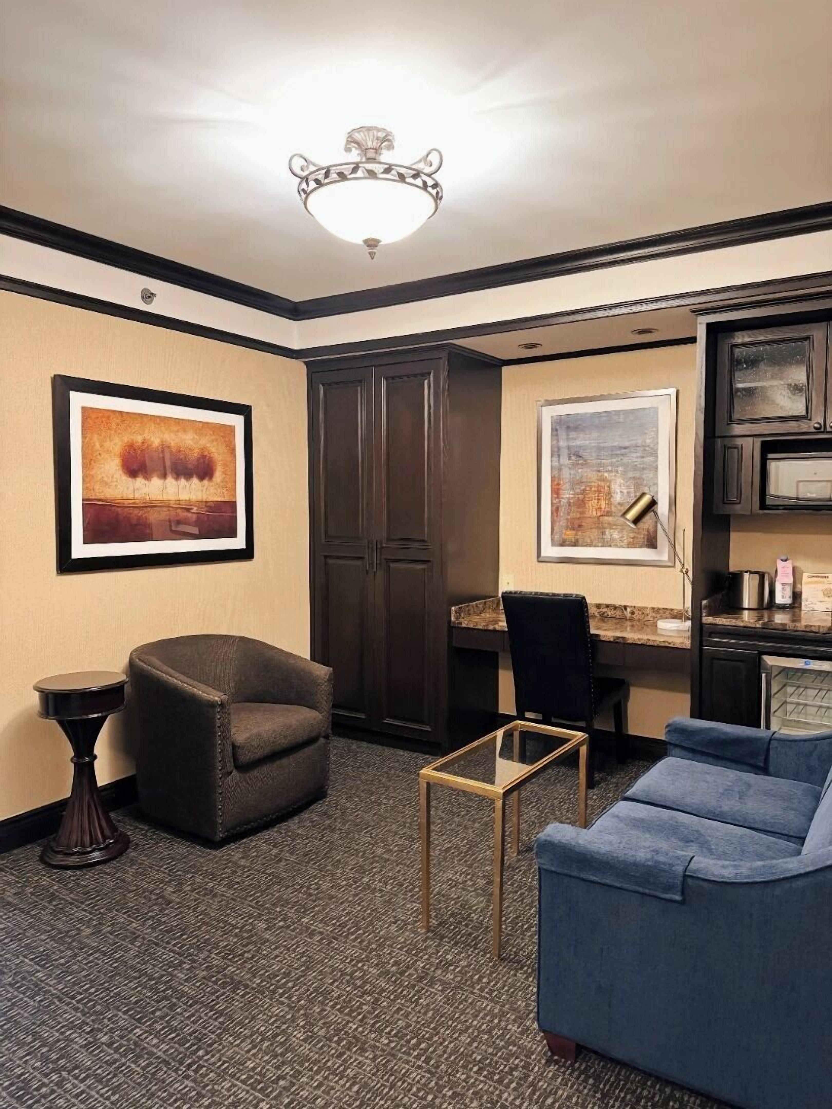 Habitación de hotel con un armario de madera oscura, un escritorio con una silla, un sofá azul, una mesita pequeña y dos obras de arte enmarcadas en las paredes.