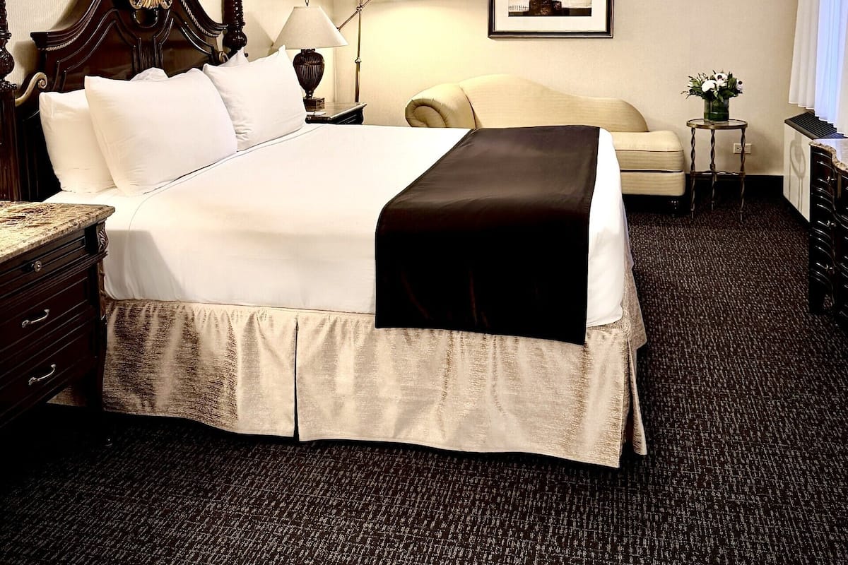 A hotel room with a large bed, a bedside table, a lamp, a framed picture on the wall, and a small table with a vase of flowers.