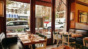 A restaurant interior with wooden tables and chairs, large windows offering a view of the street outside.
