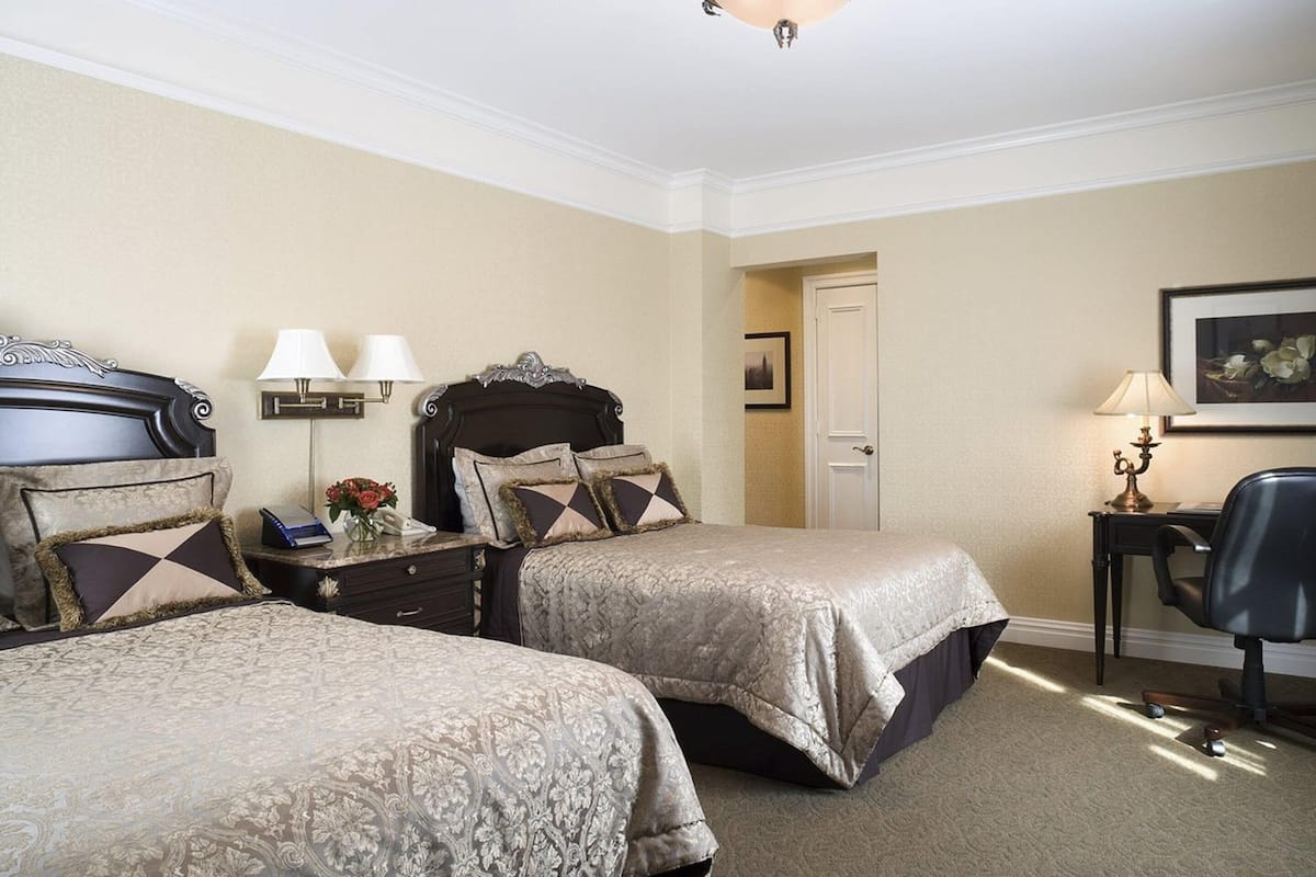 A hotel room with two beds, a desk, a chair, a lamp, and a door.