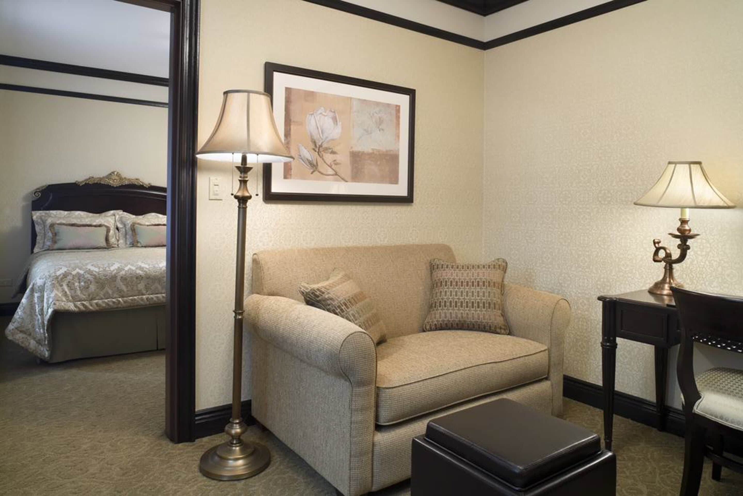 A hotel room with a beige sofa, a black ottoman, a floor lamp, a side table with a lamp, and a bed with a patterned bedspread.