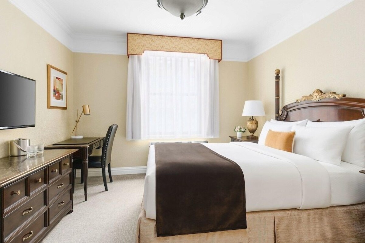 A hotel room with a bed, bedside table, lamp, chair, desk, and a television.