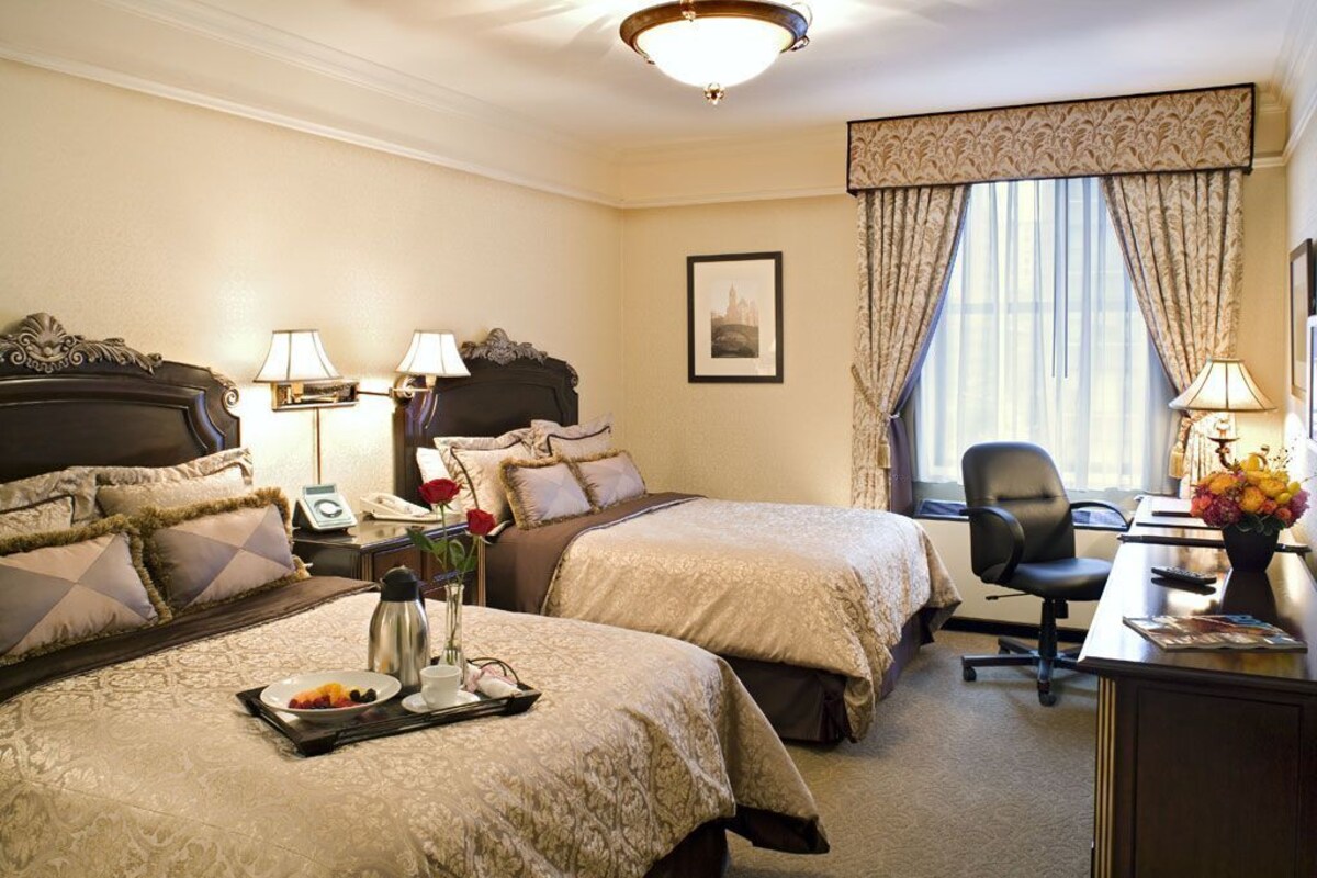 A hotel room with two beds, a desk, a chair, a window with curtains, a picture on the wall, and a vase with flowers.
