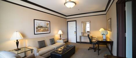 A hotel room with a sofa, armchair, ottoman, desk, chair, lamps, and a framed picture on the wall.