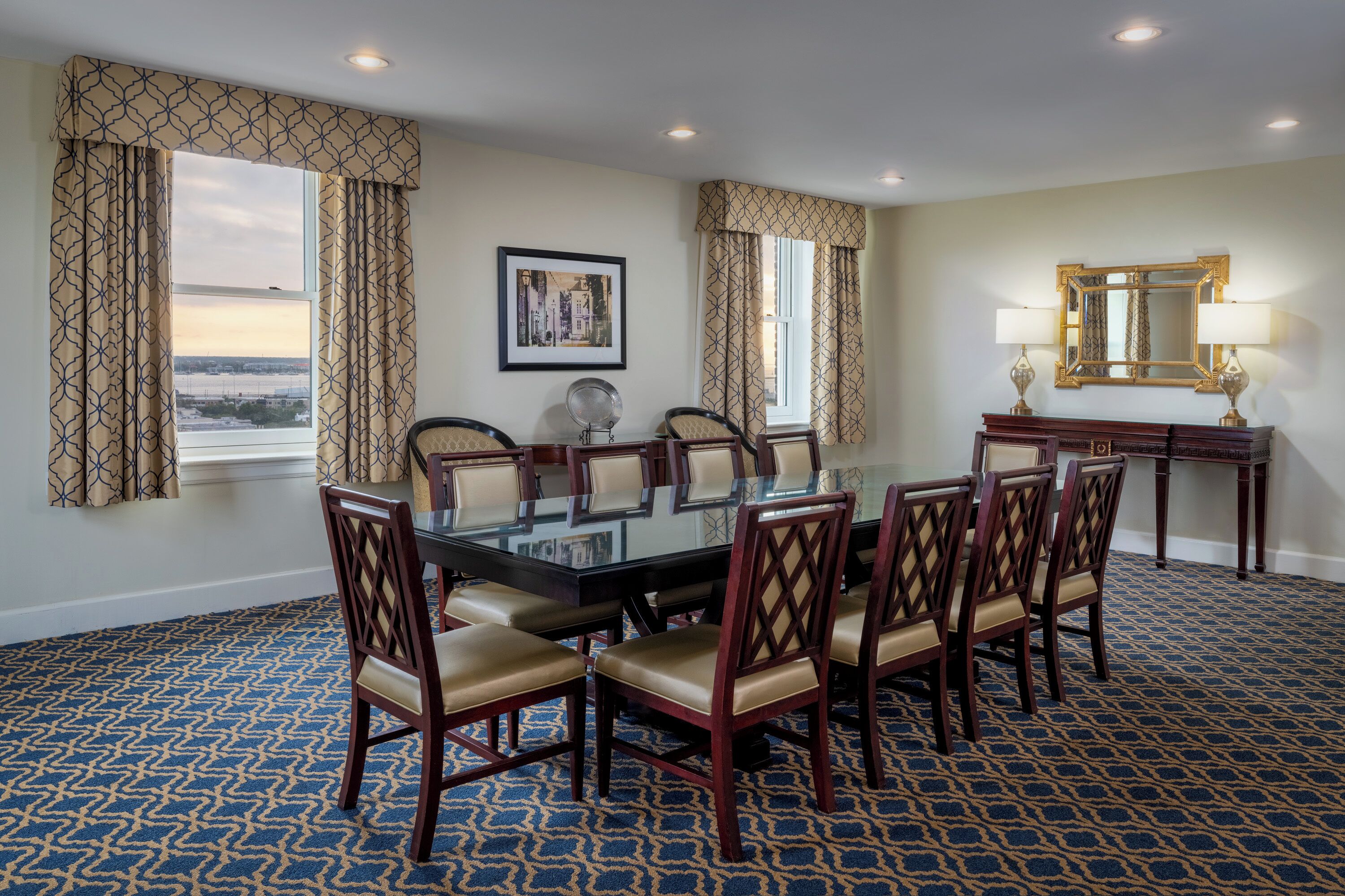 A conference room with a glass table surrounded by chairs, a framed picture on the wall, and a mirror above a desk.