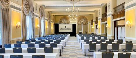 A conference room with rows of tables and chairs, a chandelier, and a screen displaying the hotel name.