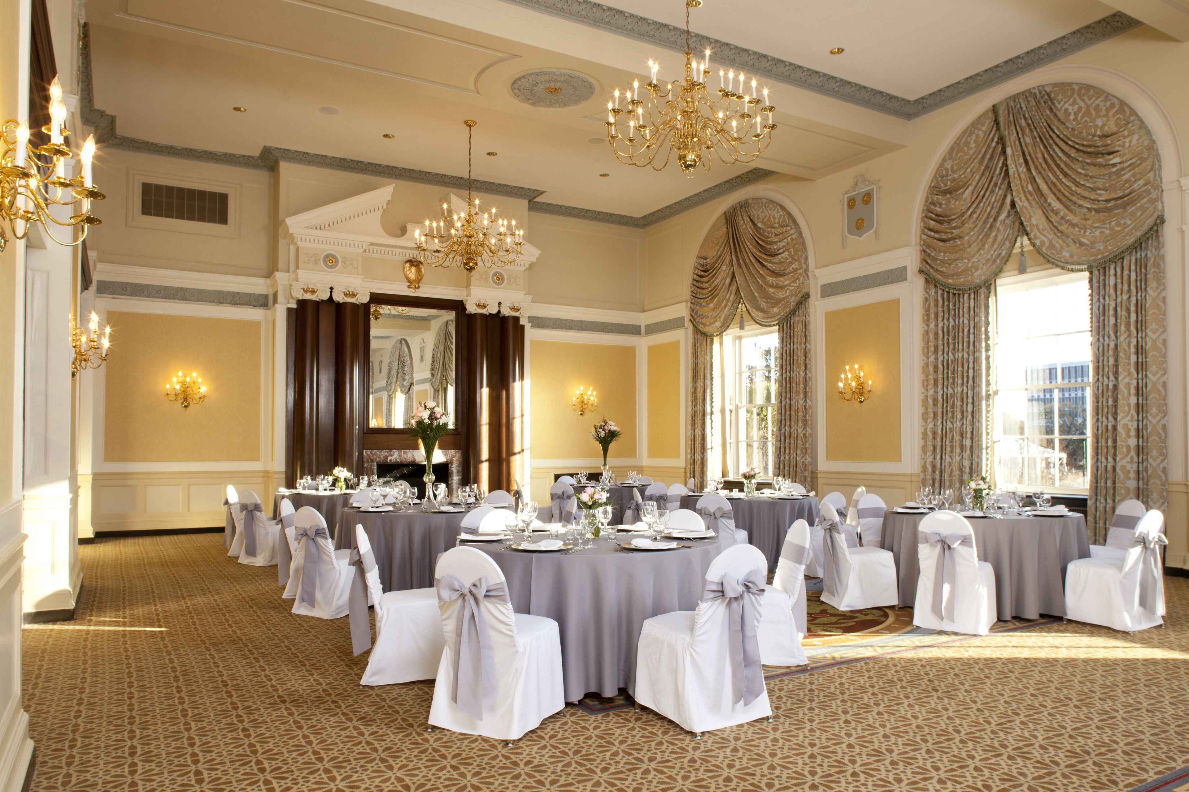 A banquet hall with round tables, white chairs, chandeliers, and large windows.