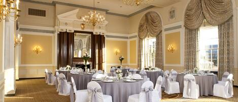 A banquet hall with round tables, white chairs, chandeliers, and large windows.