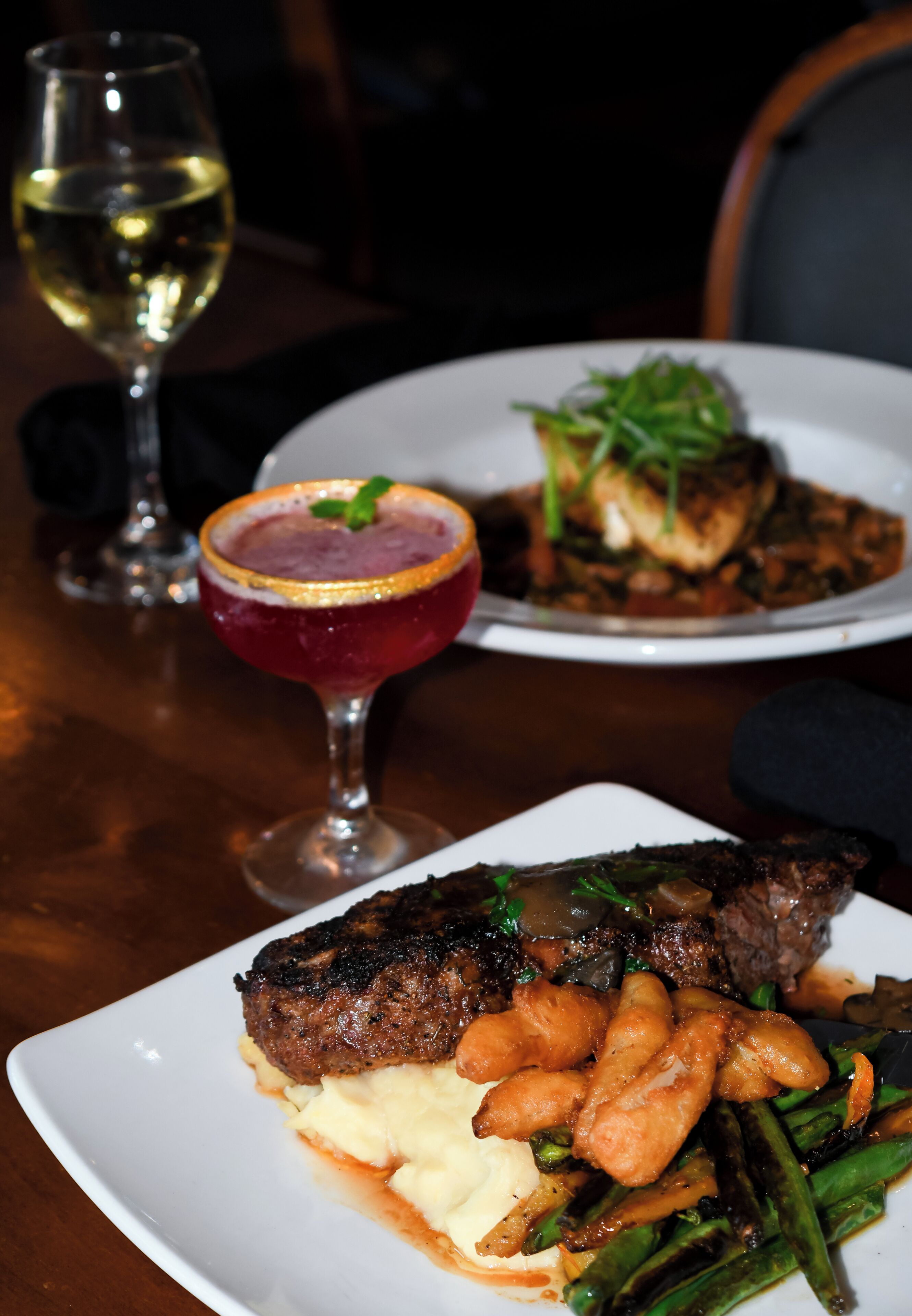 Um prato de bife com legumes e purê de batatas, uma taça de vinho branco e um coquetel com enfeite.