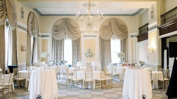 A grand ballroom with multiple round tables, elegant chandeliers, and classic drapery.