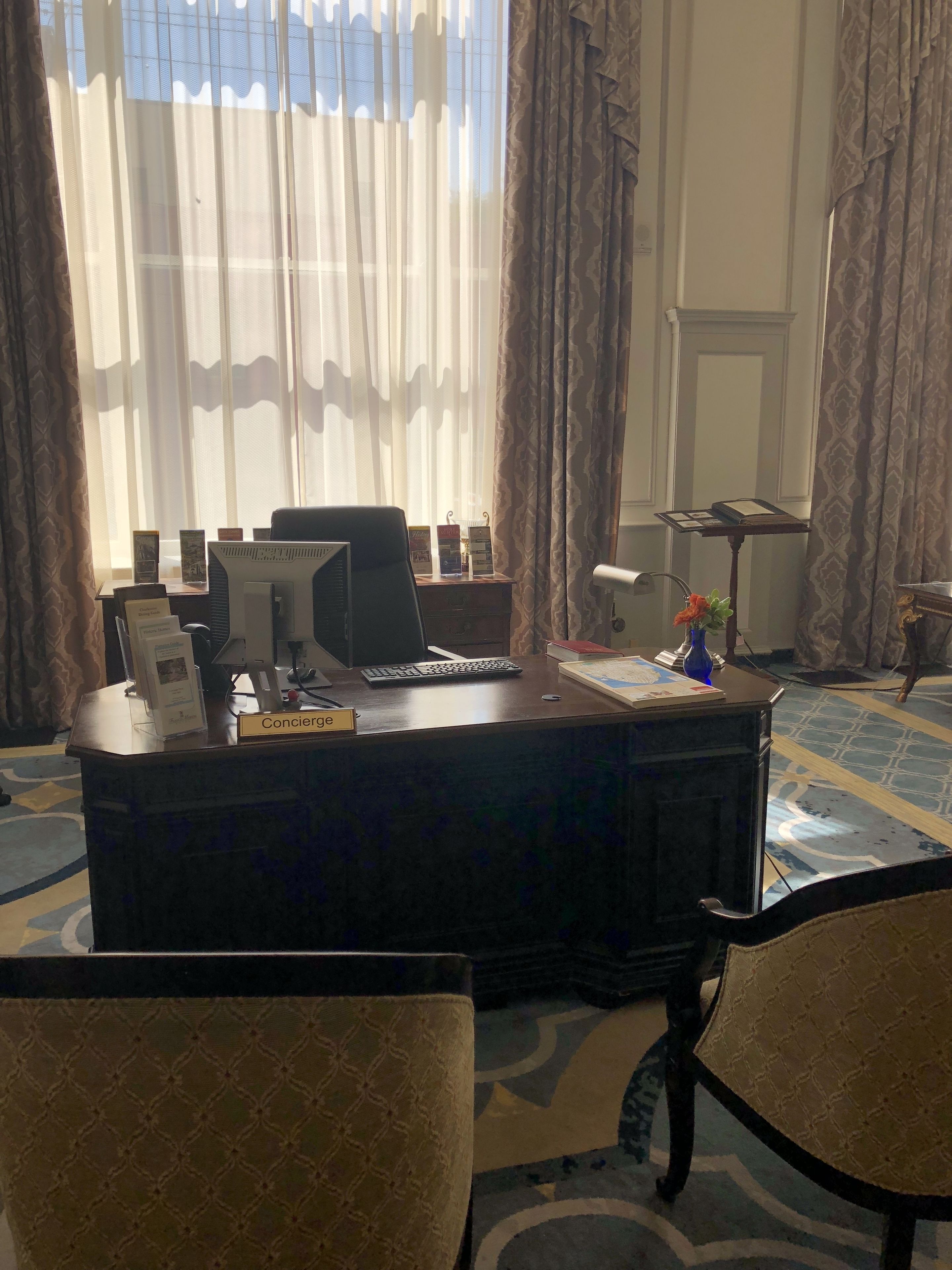 A hotel reception desk with a computer, a nameplate labeled "Concierge," and a small table with brochures. There are two chairs in front of the desk, and a window with sheer curtains allowing natural light.