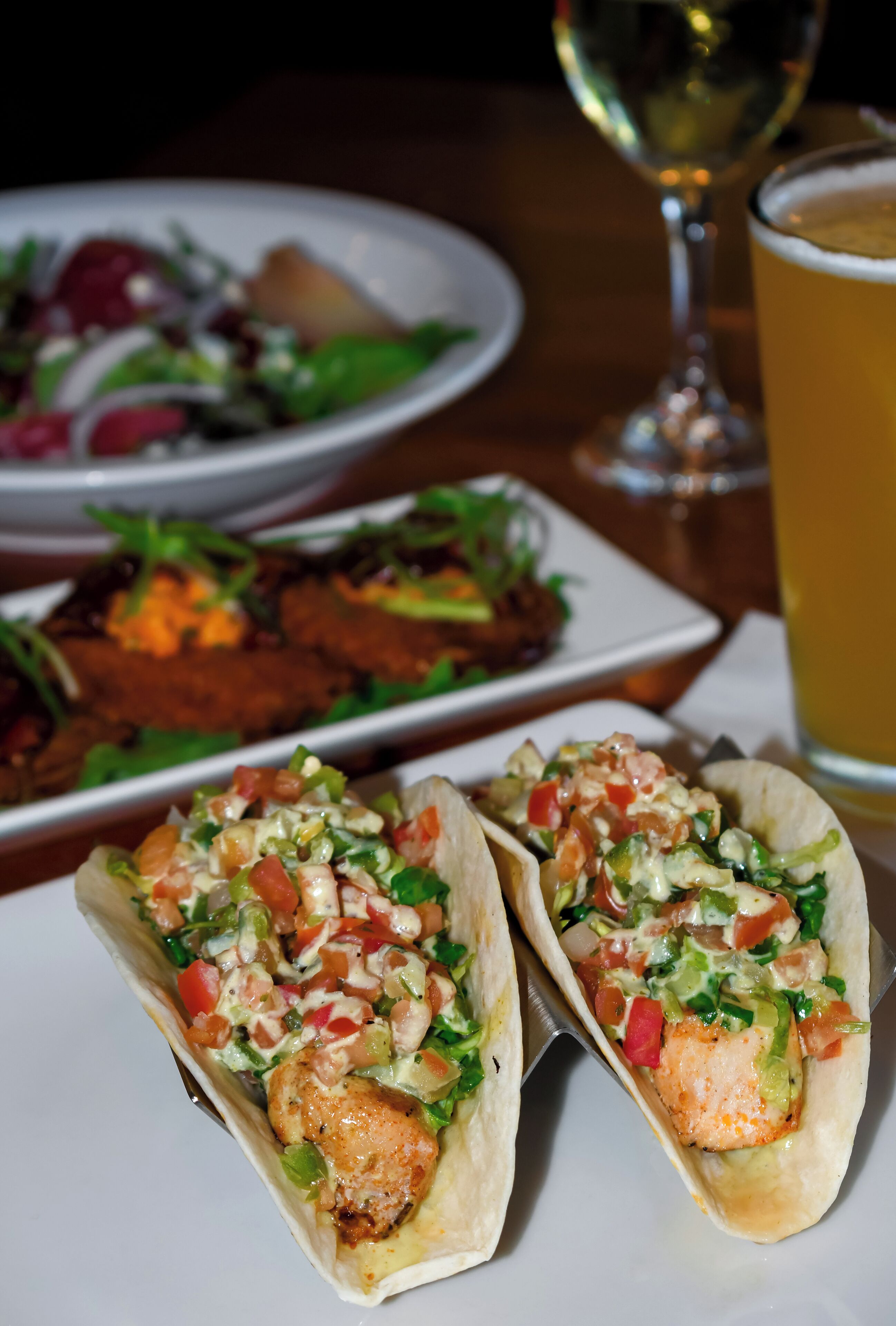 Two tacos with chicken and vegetables on a white plate, a glass of beer, and a plate of salad in the background.