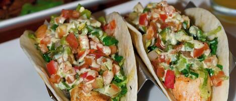 Two tacos with chicken and vegetables on a white plate, a glass of beer, and a plate of salad in the background.