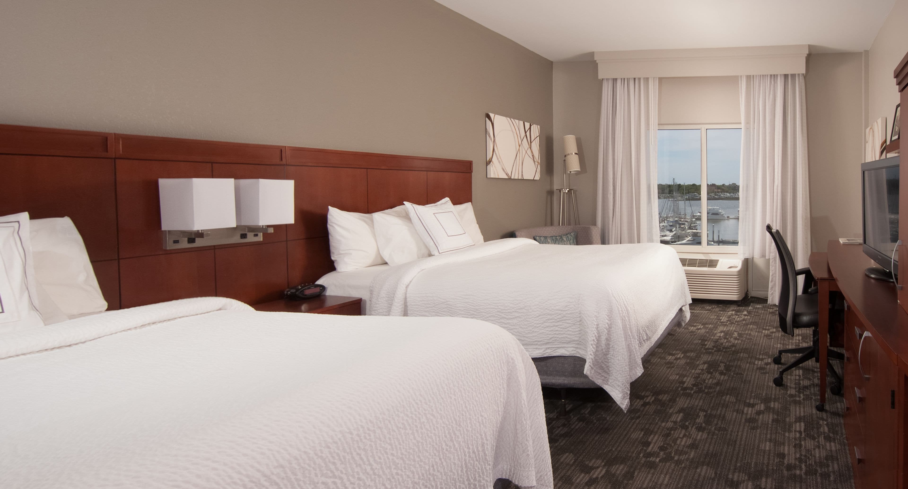 Habitación de hotel con dos camas, un escritorio, una silla, un televisor y vista al puerto deportivo.