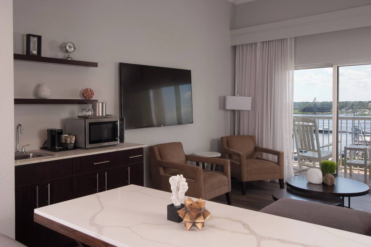 A modern hotel room with a flat-screen TV, a dining area with a marble table, two brown armchairs, a small round table with a lamp, and a view of a body of water through large windows.