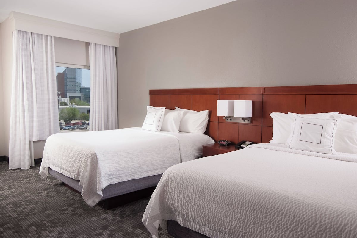 A hotel room with two beds, a wooden headboard, a nightstand, a lamp, and a view of the city through the window.