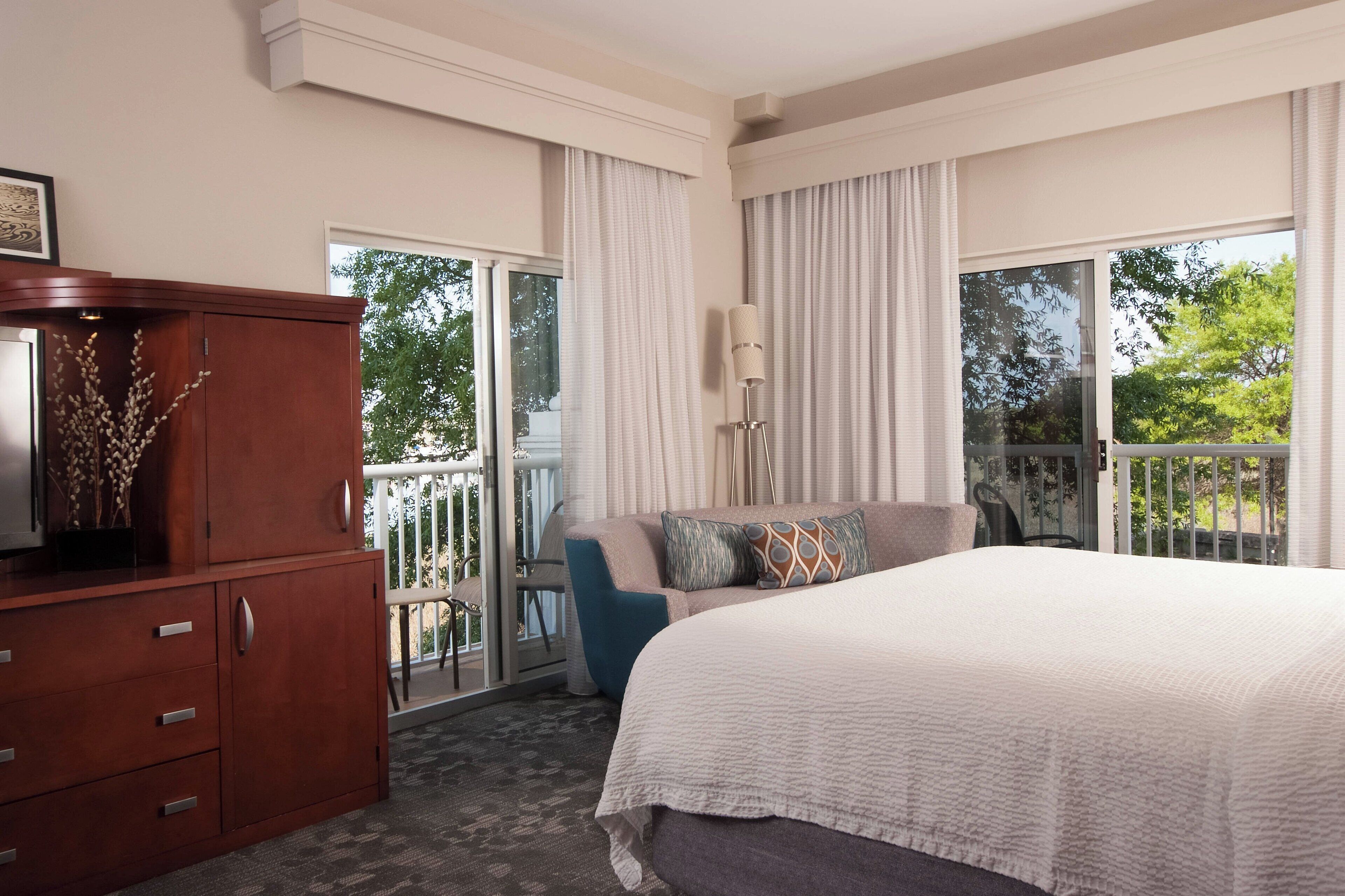 A hotel room with a large bed, a wooden dresser, a balcony with a chair, and a large window.
