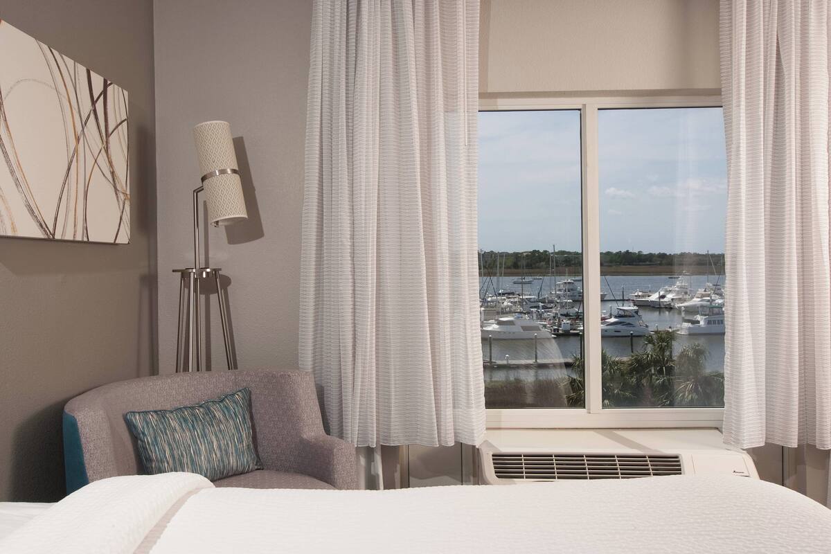 A hotel room with a bed, a chair, a floor lamp, and a large window with a marina view.