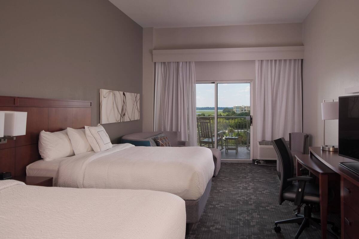 A hotel room with two beds, a desk with a TV, and a balcony with a view.