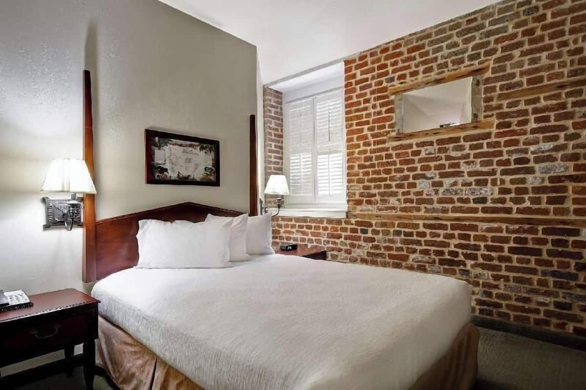Camera d'albergo con parete in mattoni, una testiera in legno, un letto con lenzuola bianche, due lampade da comodino e una finestra con persiane.