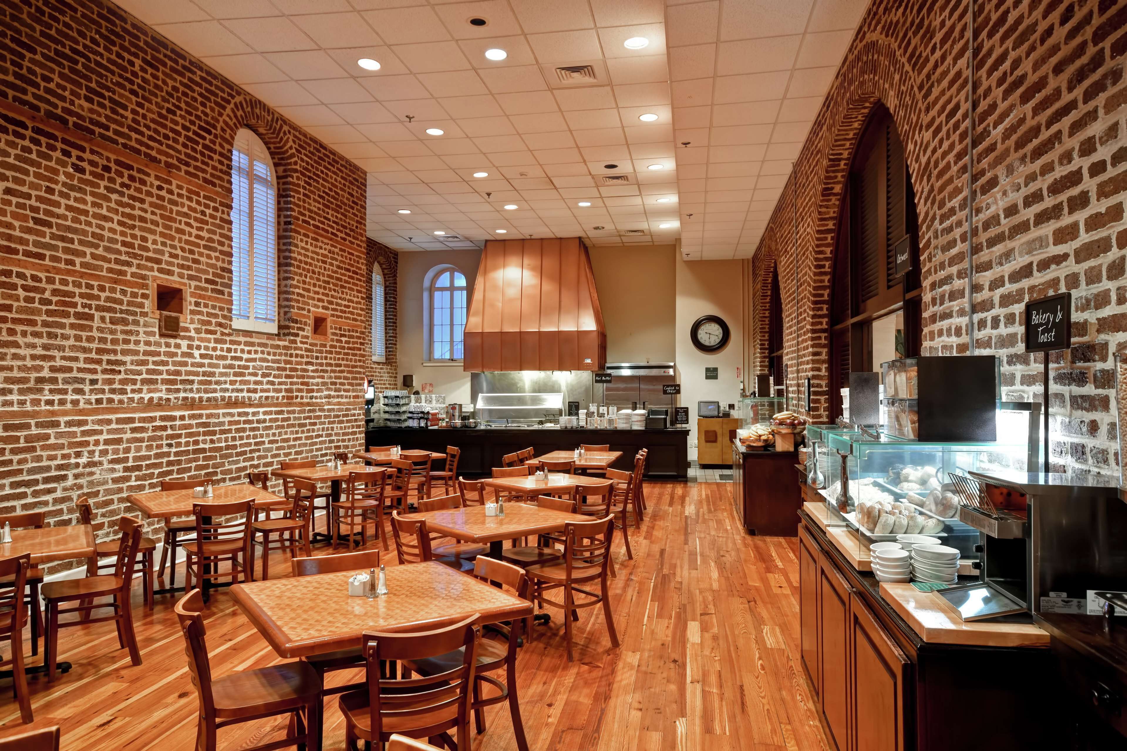 A restaurant with brick walls, wooden tables and chairs, a kitchen in the background, and a buffet counter with food items.